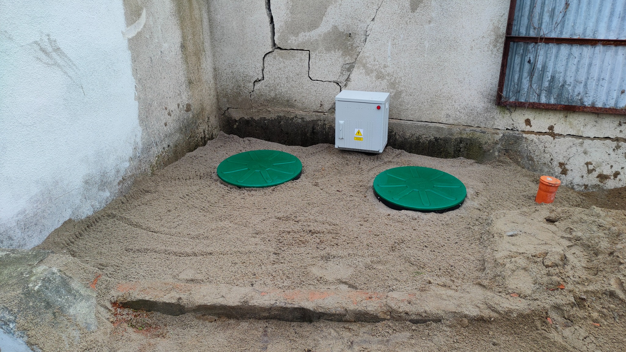 Dwa zielone włazy osadzone w piasku, widoczna skrzynka elektryczna z żółtą naklejką ostrzegawczą oraz fragment pomarańczowej rury kanalizacyjnej przy ścianie budynku z widocznymi pęknięciami.