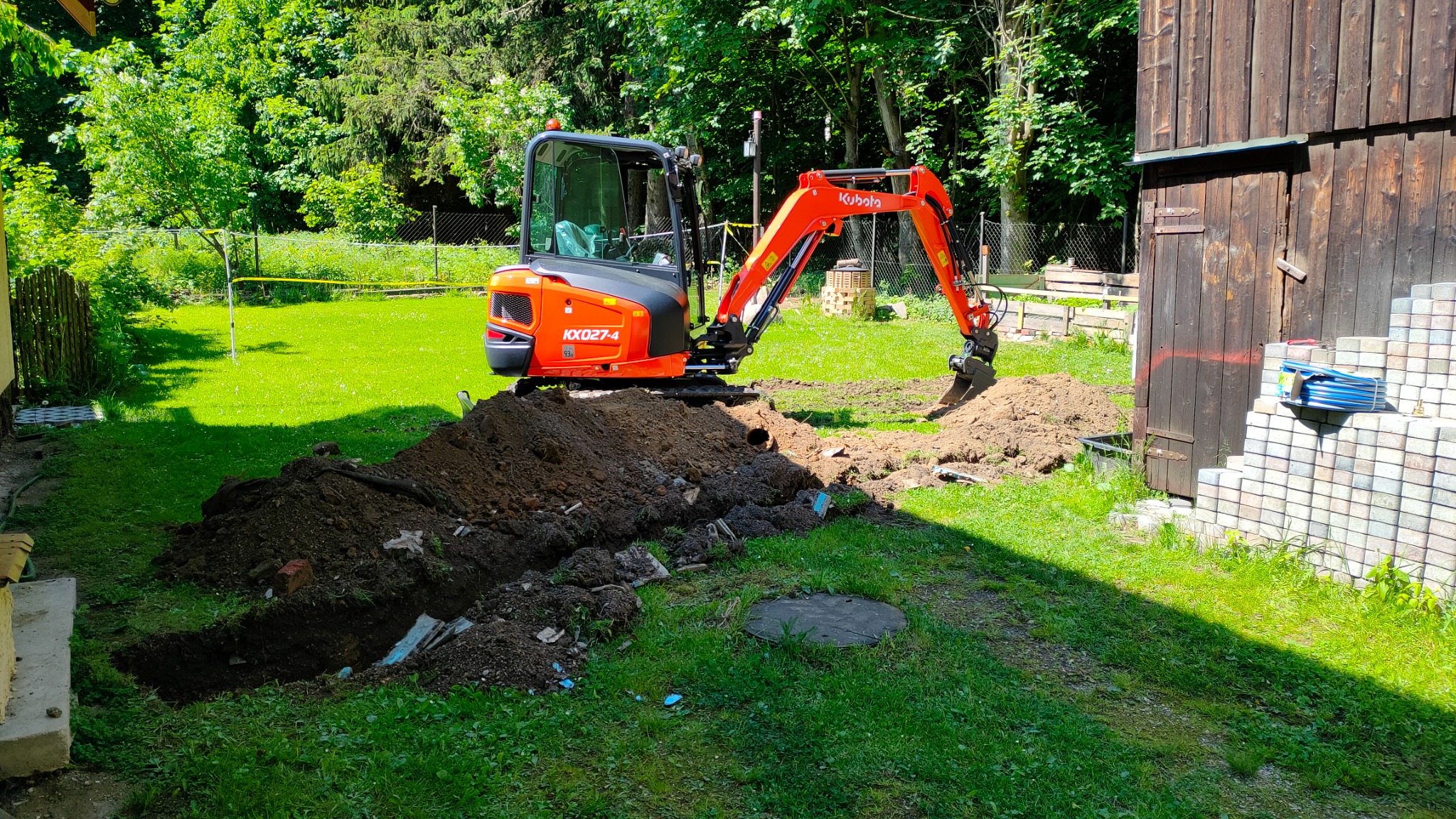 Pomarańczowa minikoparka Kubota KX027-4 wykonuje wykop w trawiastym terenie obok drewnianego budynku, widoczne ułożone kostki brukowe i zwój niebieskiego węża.