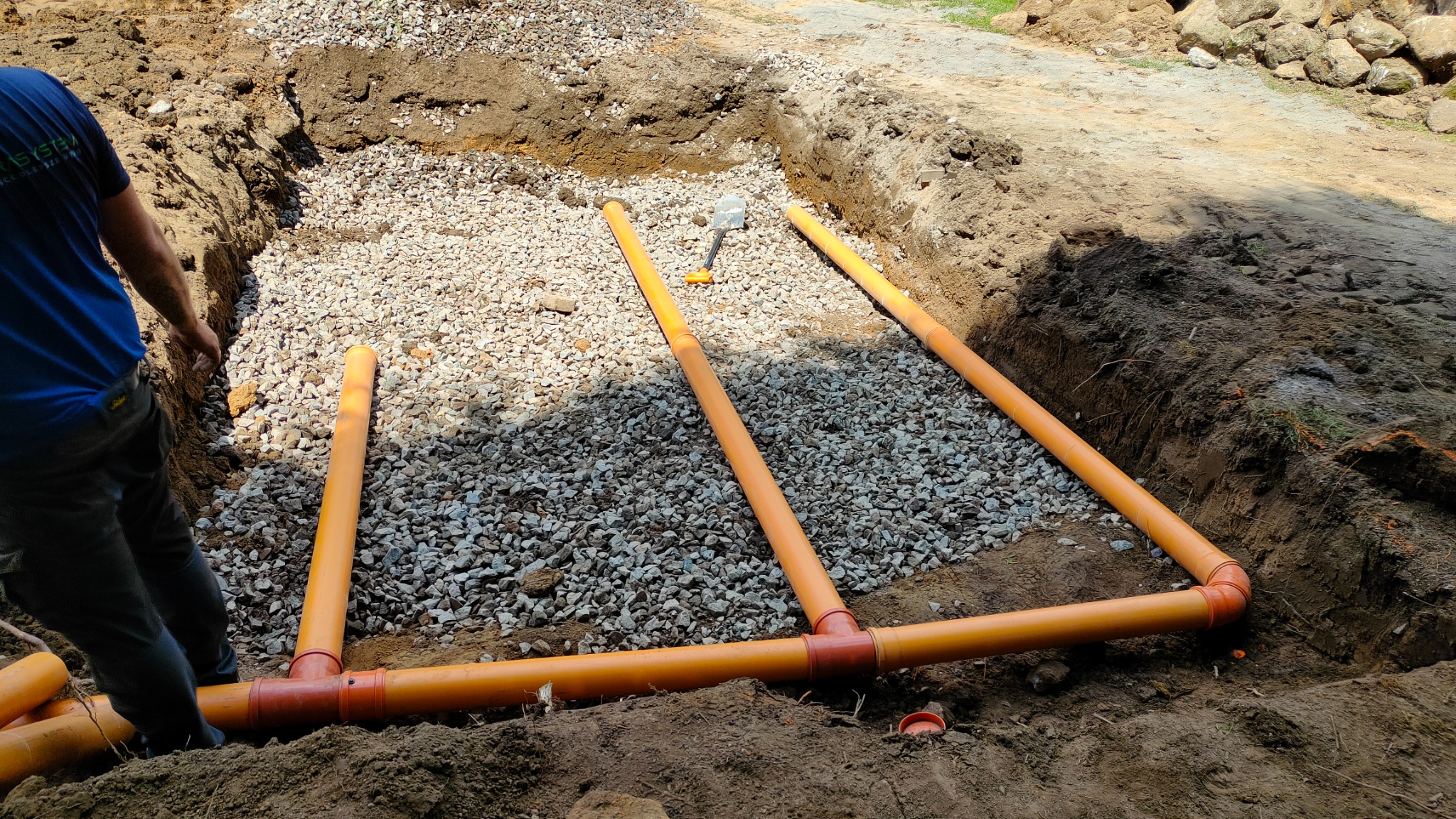 Instalacja drenażowa z pomarańczowych rur w wykopie wypełnionym żwirem, widoczna łopata i fragment sylwetki pracownika w niebieskiej koszulce.