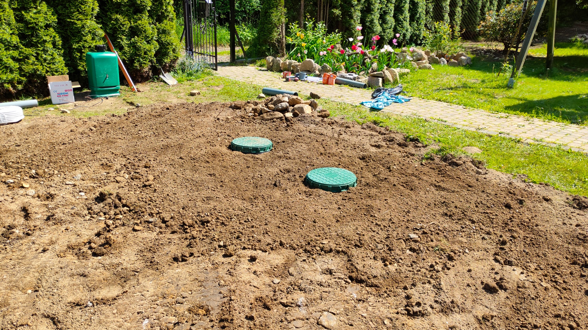 Przygotowany teren pod instalację dwóch zielonych pokryw włazów, prawdopodobnie do zbiorników, z widocznym wykopem, narzędziami i elementami instalacji w tle, na tle ogrodu z kwitnącymi tulipanami.