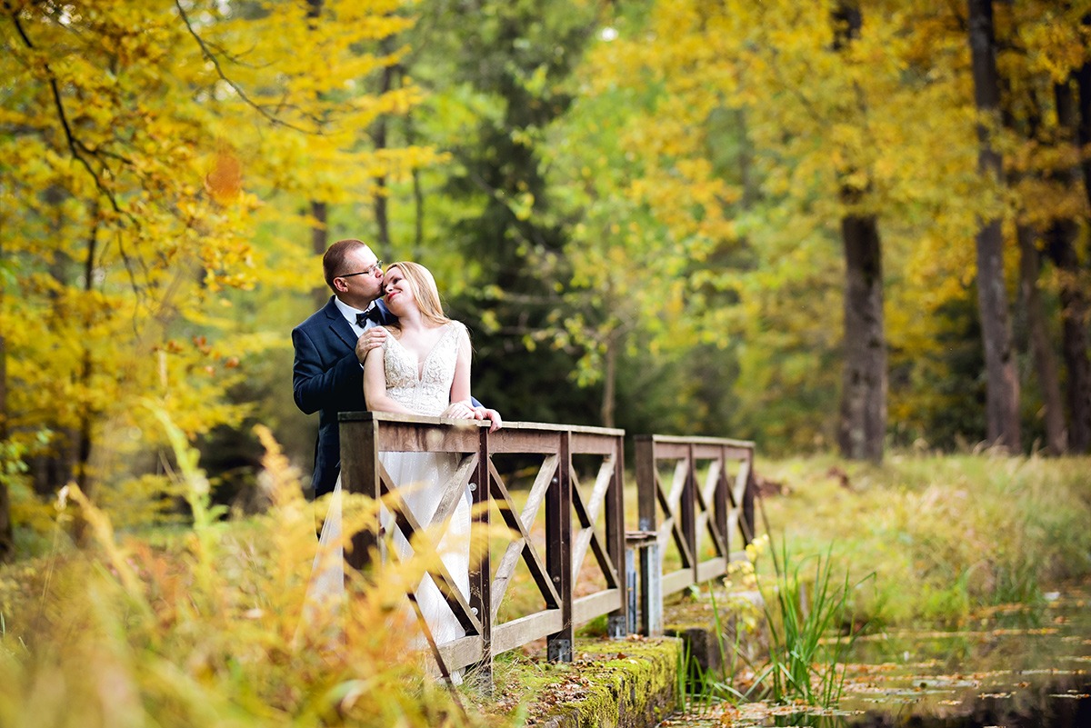 Jesienny plener ślubny: Pan młody całuje pannę młodą w czoło na drewnianym mostku z widokiem na park z żółtymi drzewami.