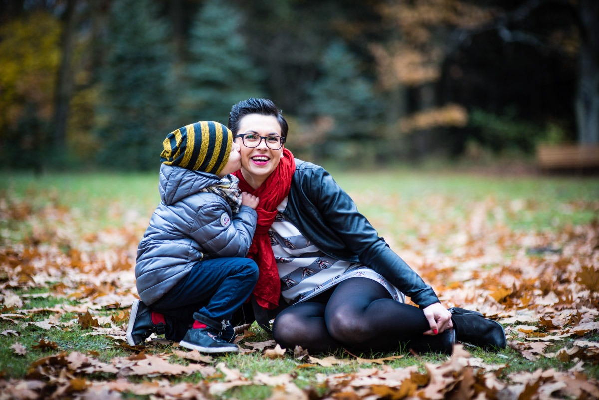 Uśmiechnięta kobieta w okularach siedzi na trawie w parku, a mały chłopiec w czapce w paski całuje ją w policzek. W tle jesienne drzewa i opadłe liście.