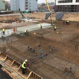 Wykonywanie fundamentów żelbetowych: widok z góry na zbrojenie i wylewanie betonu, pracownicy w kamizelkach odblaskowych, niebieskie rury wystające z gruntu, dźwig w tle.