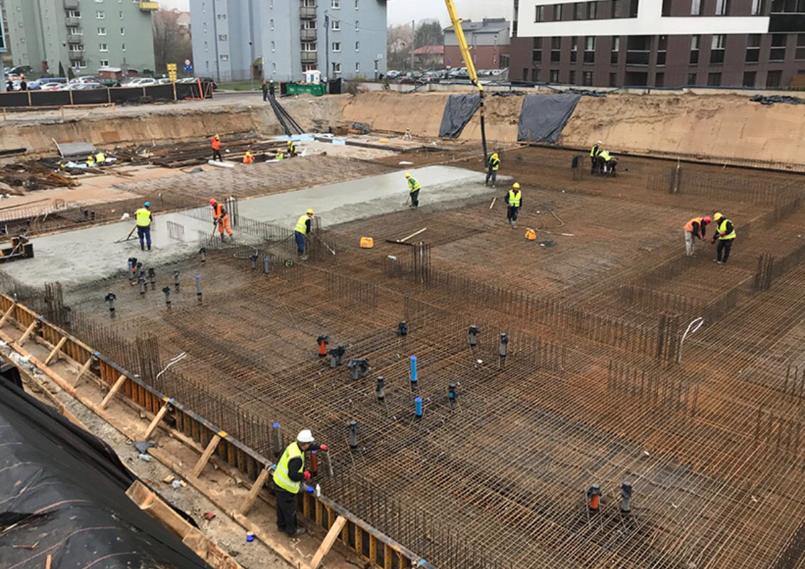 Wykonywanie fundamentów żelbetowych: widok z góry na zbrojenie i wylewanie betonu, pracownicy w kamizelkach odblaskowych, niebieskie rury wystające z gruntu, dźwig w tle.