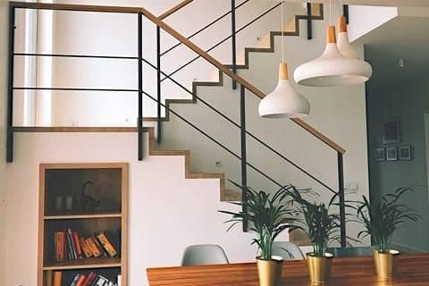 Wewnętrzne schody z drewnianymi stopniami i czarną, minimalistyczną balustradą, oświetlone wiszącymi lampami w kształcie dzwonków, uzupełnione roślinami w złotych doniczkach i regałem z książkami.