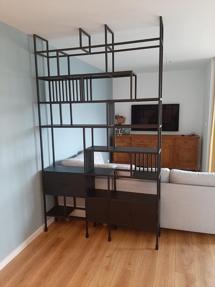 Metalowy regał ażurowy w stylu industrialnym, z czarną ramą i półkami, stojący w salonie na drewnianej podłodze, widoczny fragment sofy i komody w tle.