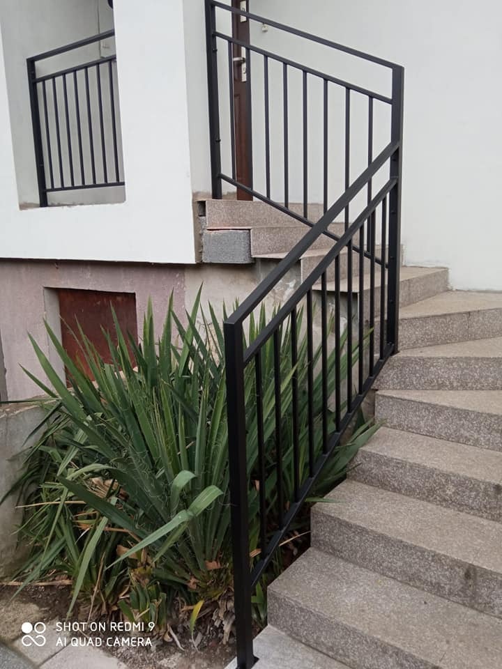 Czarna metalowa balustrada zewnętrzna przy schodach wejściowych do domu, z roślinnością u podstawy.