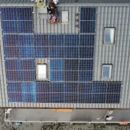 Widok z góry na instalację paneli słonecznych na dachu z szarej dachówki, biały bus zaparkowany obok budynku, dwóch instalatorów na dachu, jeden na drabinie, drugi montuje panele.