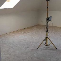 Poddasze ze skośnym sufitem i oknem dachowym, ściany przygotowane do malowania, widoczny parkiet i lampa budowlana na statywie.