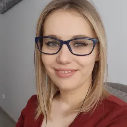 Portret uśmiechniętej blondynki w okularach, pracującej zdalnie w Kielcach, z widocznym obrazem na ścianie w tle.