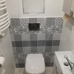 Biała toaleta i umywalka w małej łazience z szaro-białymi wzorzystymi płytkami na ścianie, widoczny grzejnik drabinkowy i podajnik na papier toaletowy.