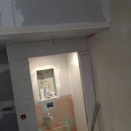 Uslugi remontowe - Łazienka w trakcie prac: zamontowana toaleta, ściany przygotowane pod płytki, zabudowa z płyty karton gips z wnęką na kosmetyki, widoczne instalacje elektryczne.
