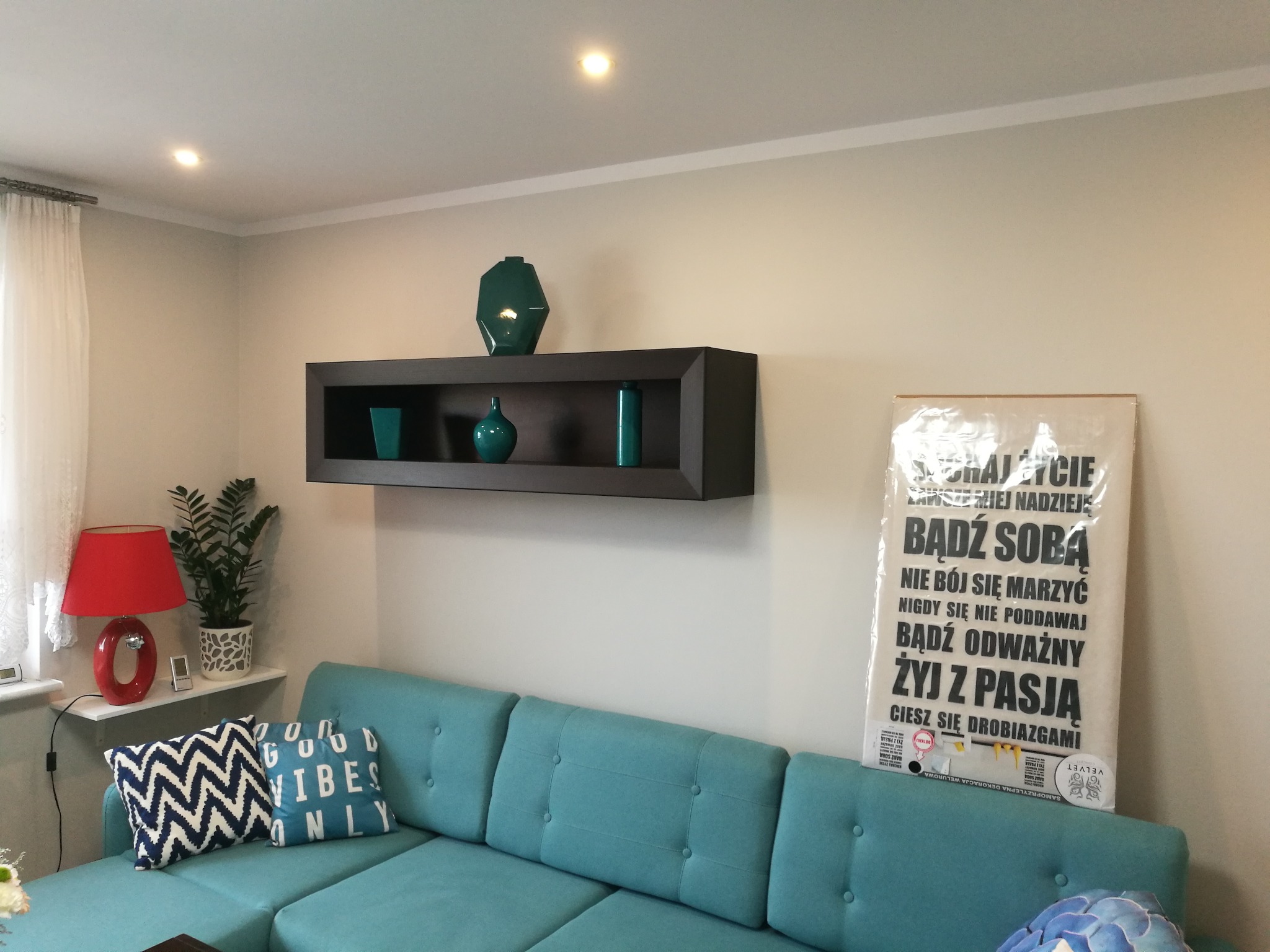 Turkusowa sofa z poduszkami w salonie, nad nią półka z dekoracyjnymi wazonami, plakat z napisem 'Żyj z pasją' i lampa z rośliną na parapecie.