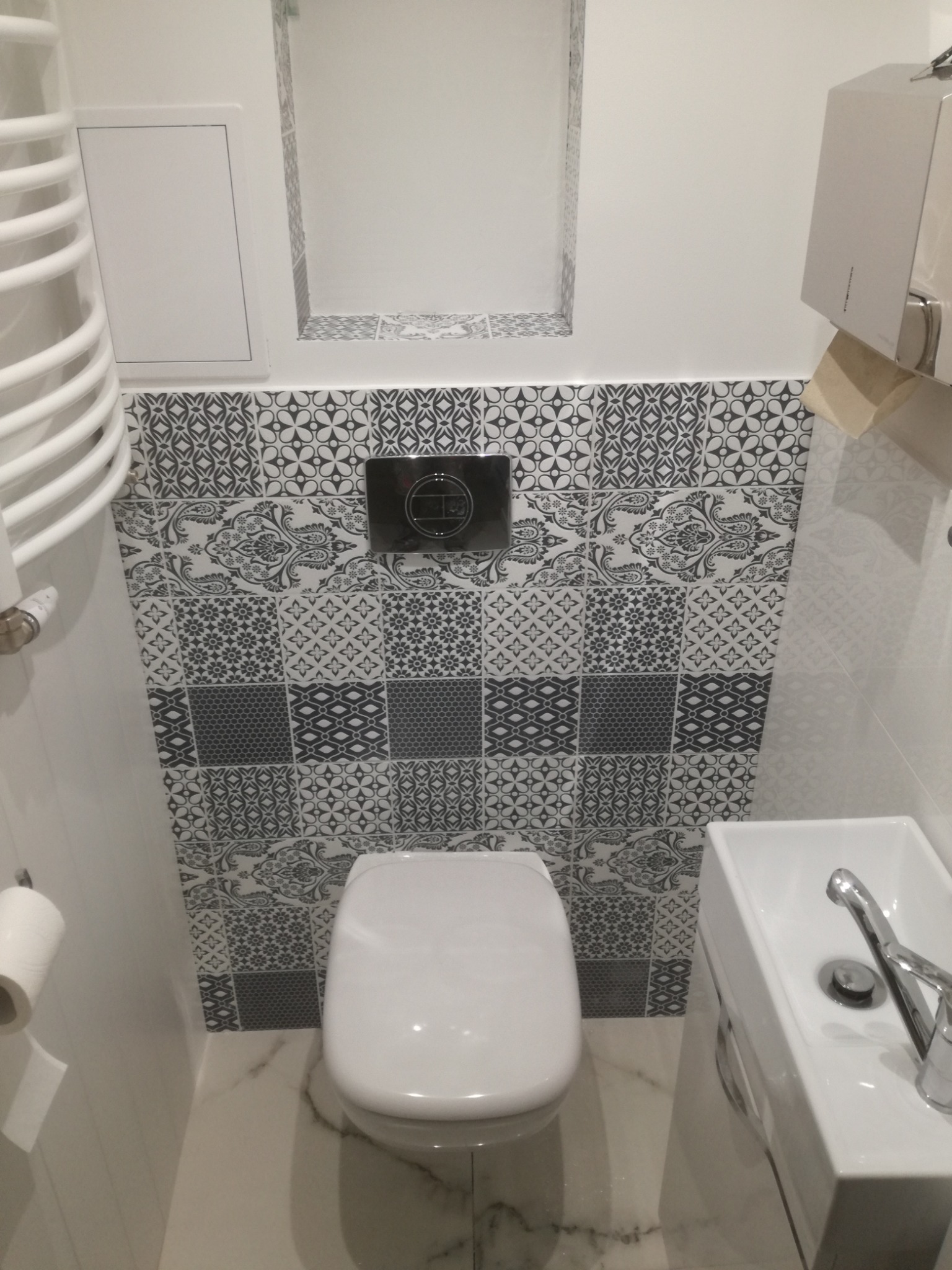 Biała toaleta i umywalka w małej łazience z szaro-białymi wzorzystymi płytkami na ścianie, widoczny grzejnik drabinkowy i podajnik na papier toaletowy.