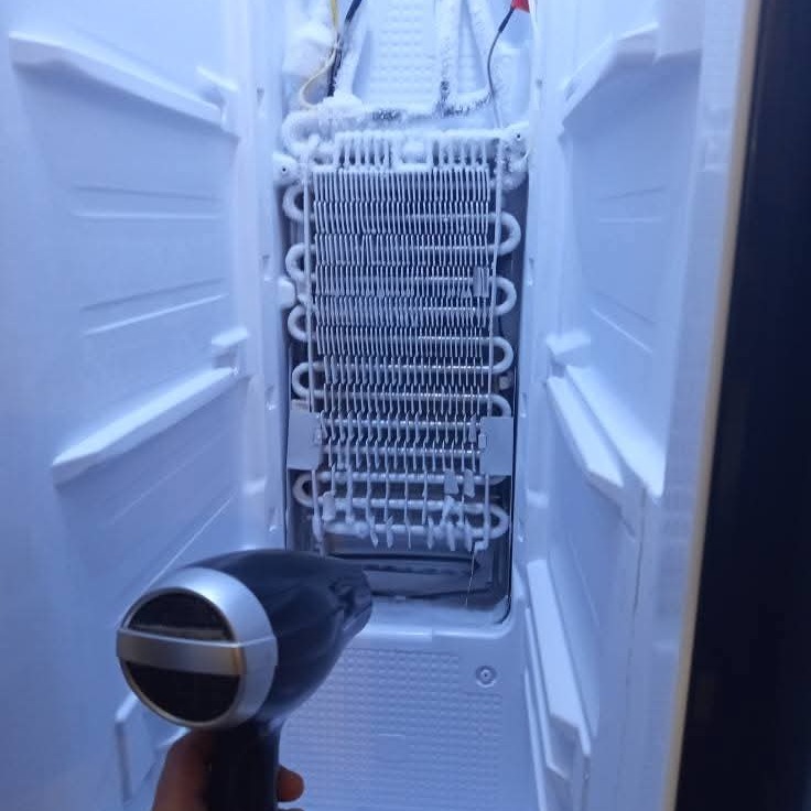 Odszranianie zamrażarki suszarką do włosów. Widoczne zamrożone elementy chłodzące wewnątrz otwartej komory. Nietypowy sposób na szybkie usunięcie lodu.
