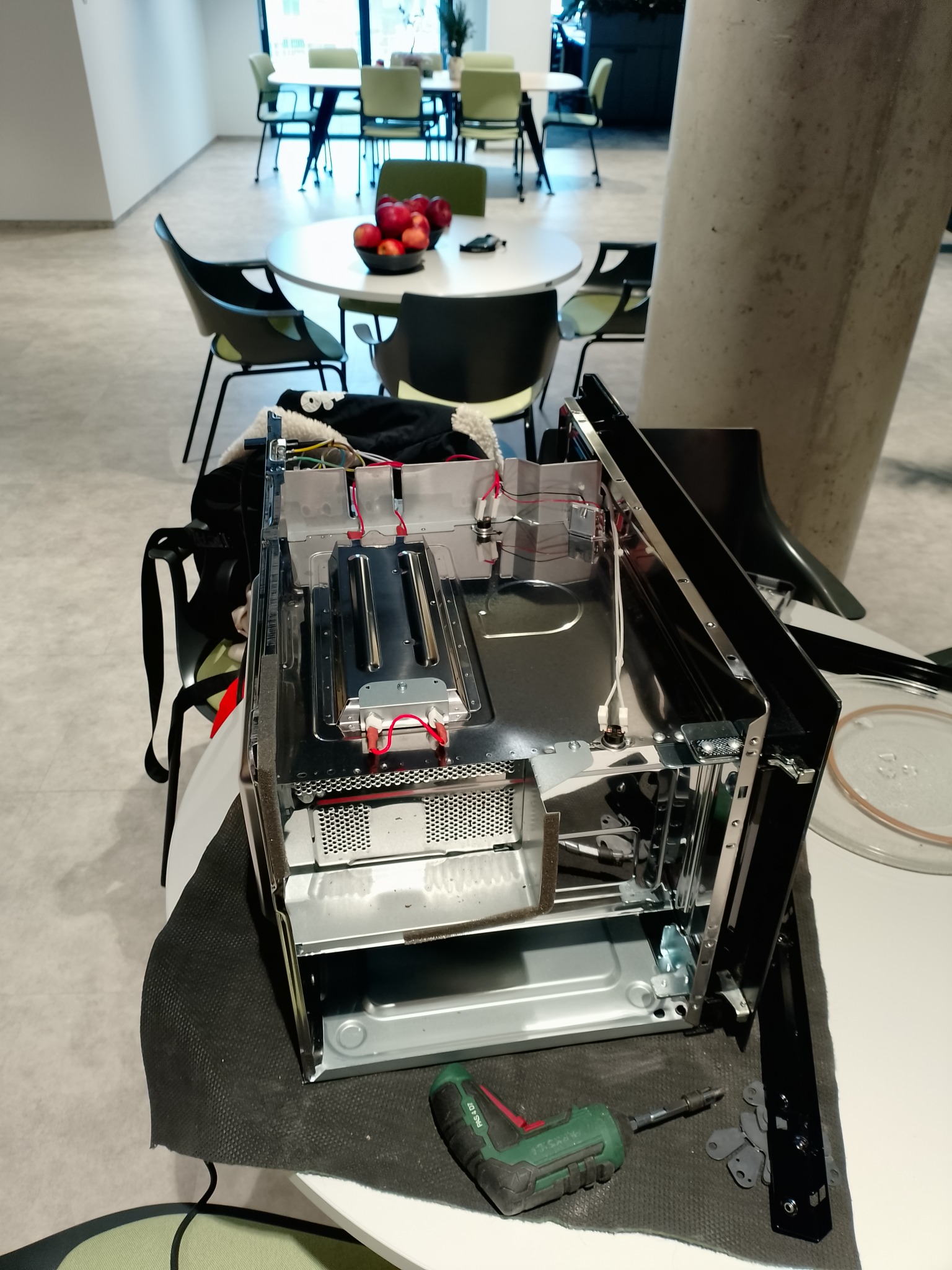 Rozłożona kuchenka mikrofalowa na stole w jasnym pomieszczeniu, widoczne elementy wewnętrzne i narzędzie. W tle stoliki i krzesła.