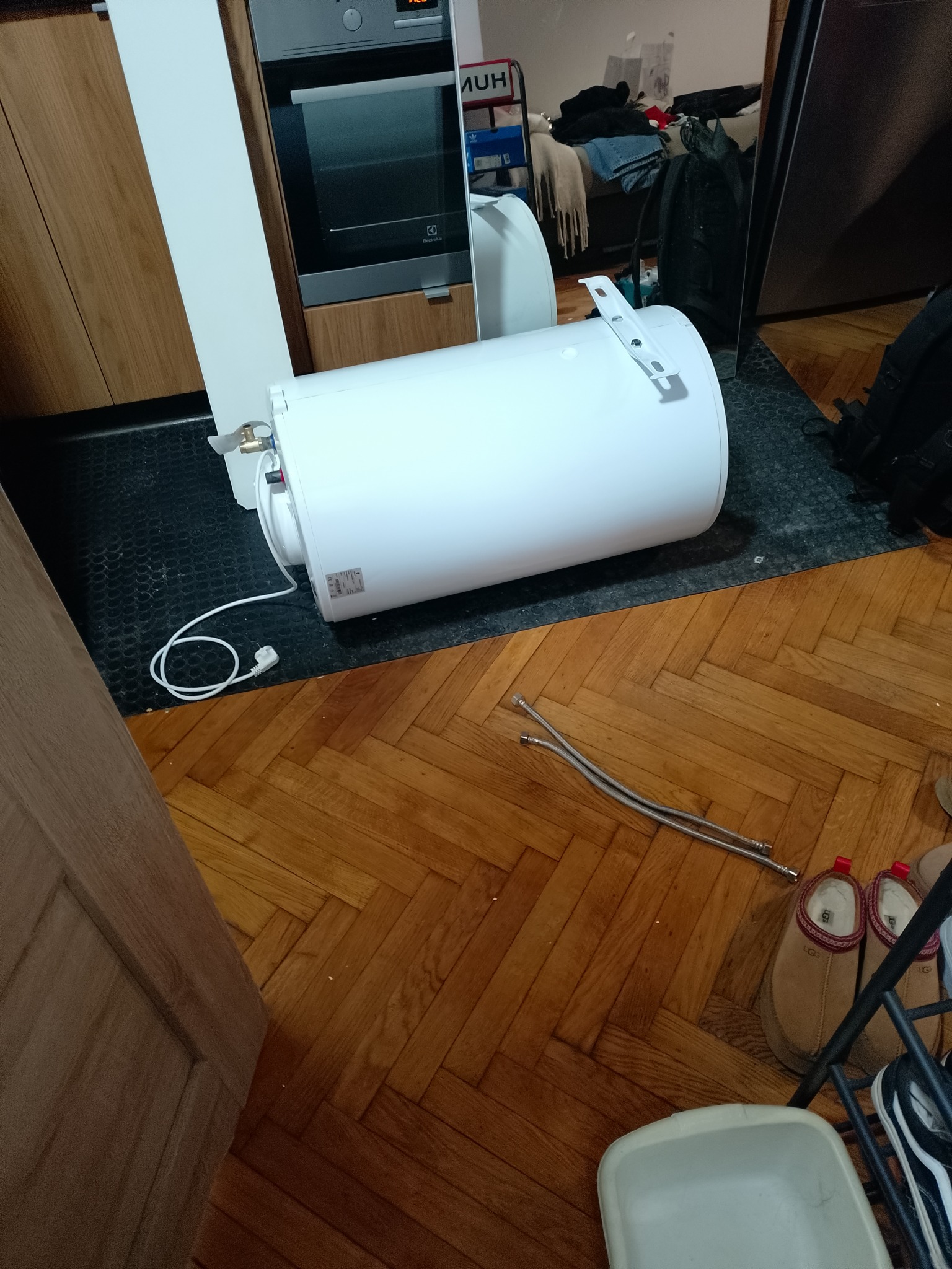 Biały, poziomy bojler elektryczny z kablem i rurkami przyłączeniowymi leży na podłodze w kuchni, obok zabudowy i piekarnika Electrolux. Widoczne akcesoria montażowe.