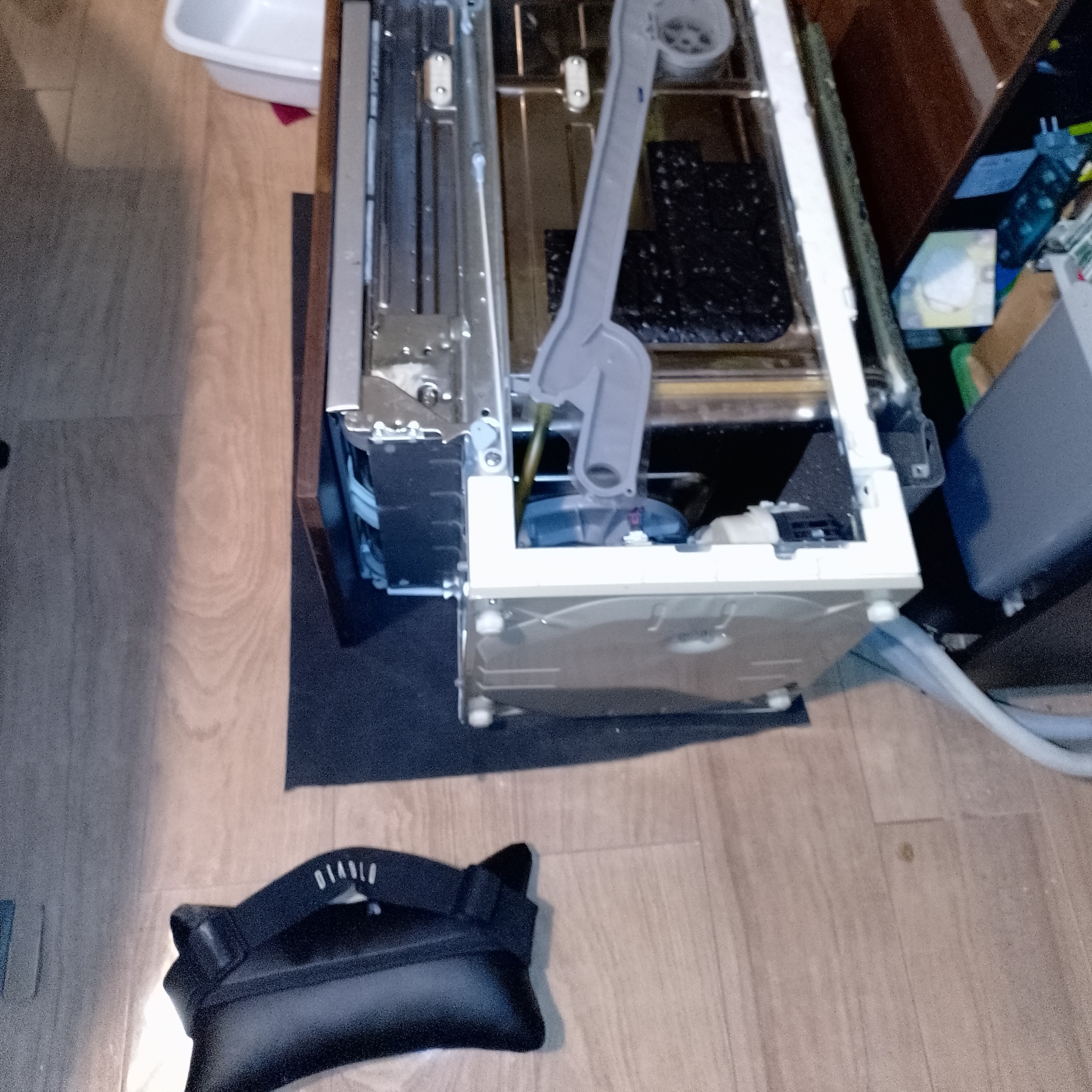 Rozłożona zmywarka na podłodze z widocznymi elementami wewnętrznymi, gotowa do naprawy. Na pierwszym planie leży czarna saszetka.