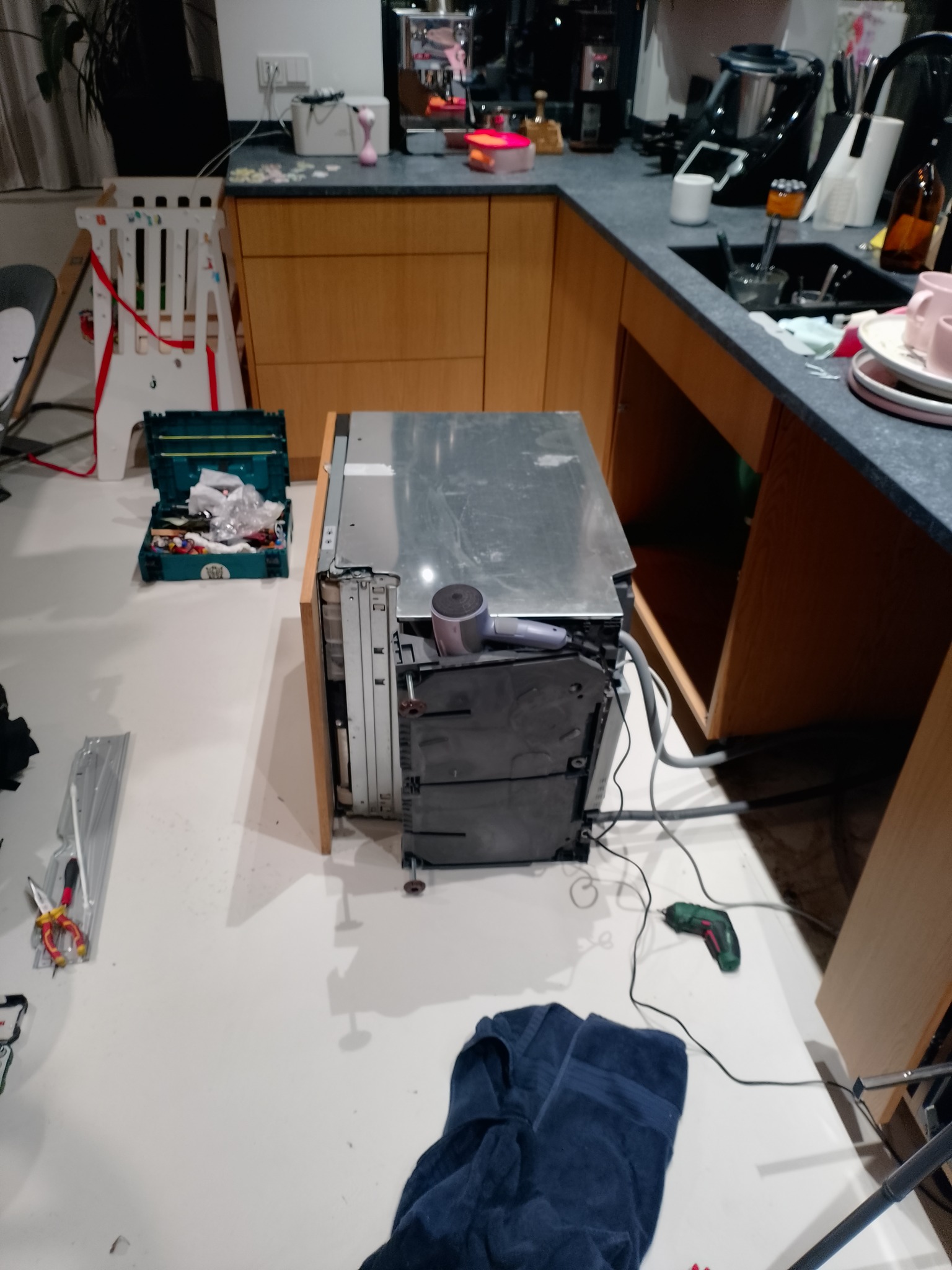 Rozmontowana zmywarka na podłodze w kuchni, obok skrzynka z narzędziami i wkrętarka. Widoczne elementy instalacji i wnętrze urządzenia.