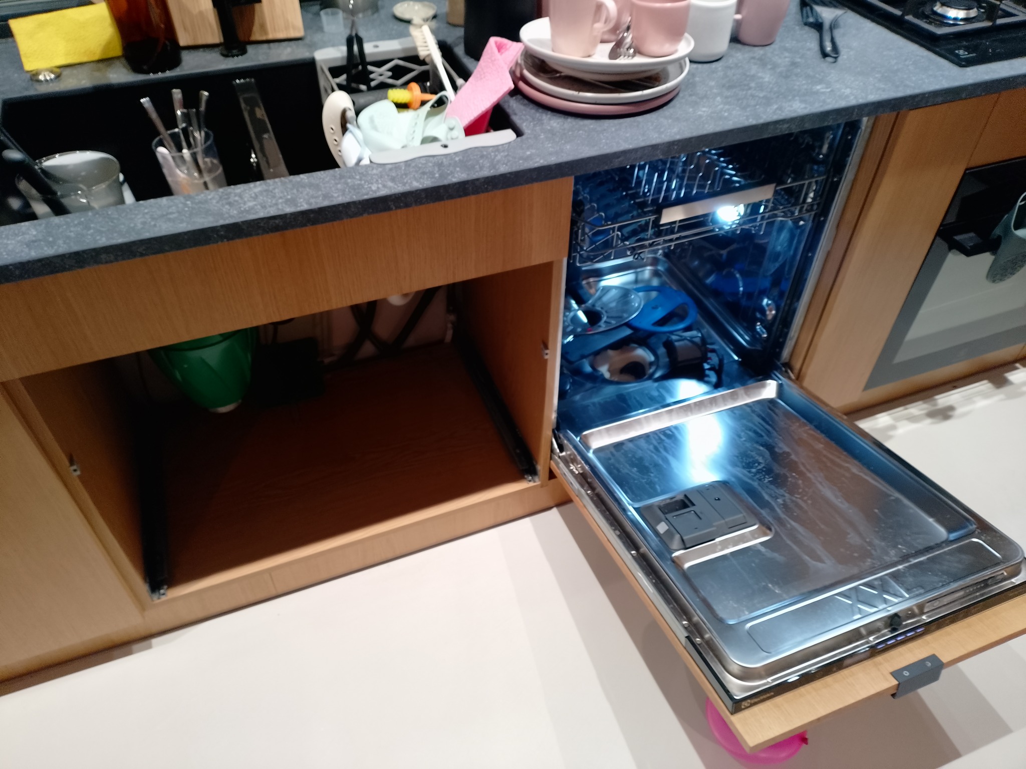 Otwarta zmywarka zabudowana w szafce kuchennej, widoczne naczynia w środku, obok zlewozmywak z naczyniami i akcesoriami kuchennymi. Oświetlenie wnętrza zmywarki.