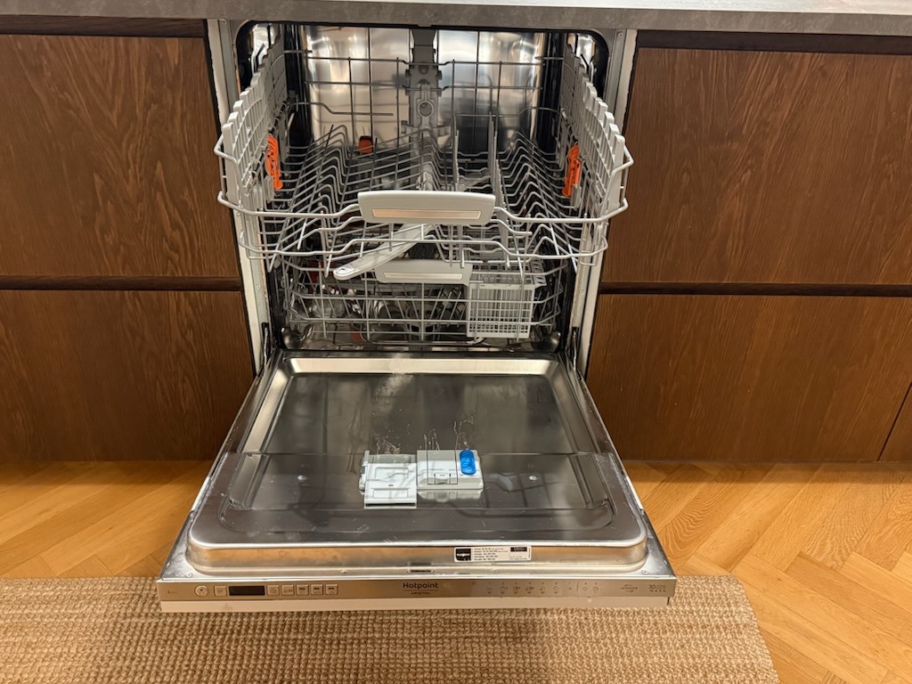 Otwarta zmywarka Hotpoint z widocznymi koszami i wnętrzem, wbudowana w drewnianą zabudowę kuchenną, na tle parkietu i dywanu.