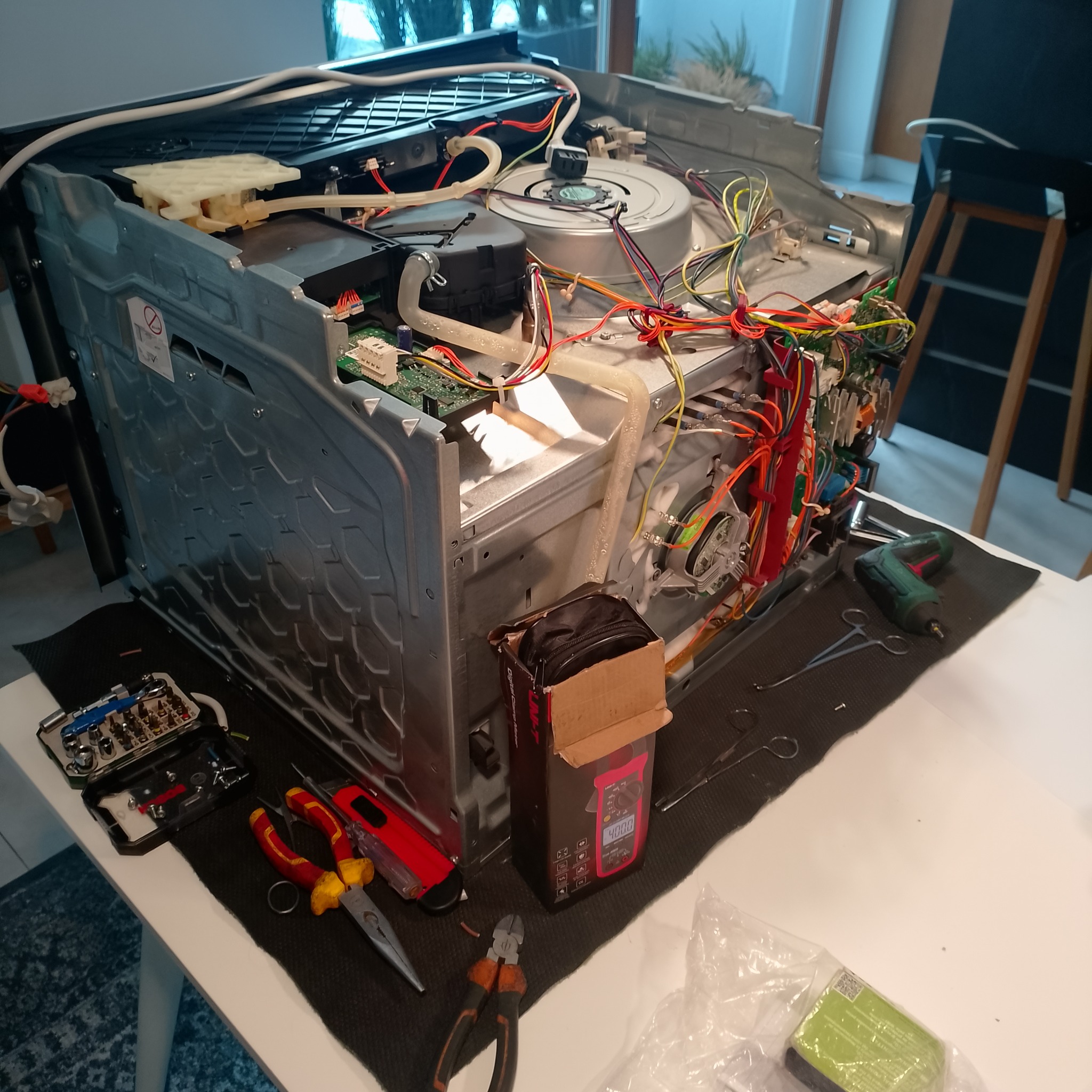 Rozłożona zmywarka na stole z narzędziami: śrubokręty, kombinerki, miernik. Widoczne wnętrze urządzenia z przewodami i elementami elektronicznymi.