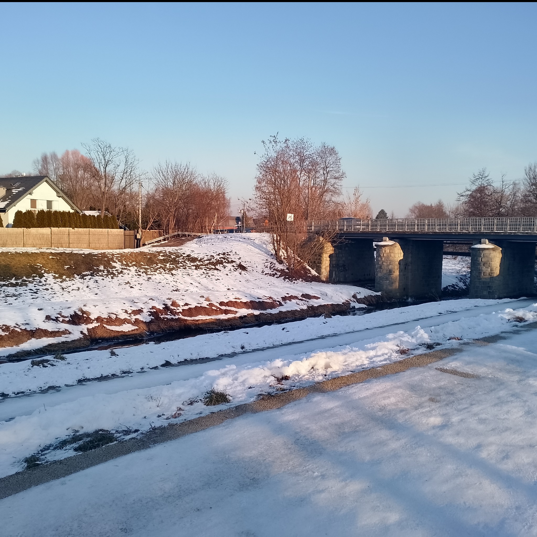 Zimowy krajobraz z rzeką, mostem i domem w tle, oświetlony popołudniowym słońcem. Śnieg pokrywa brzegi rzeki i okoliczne tereny.