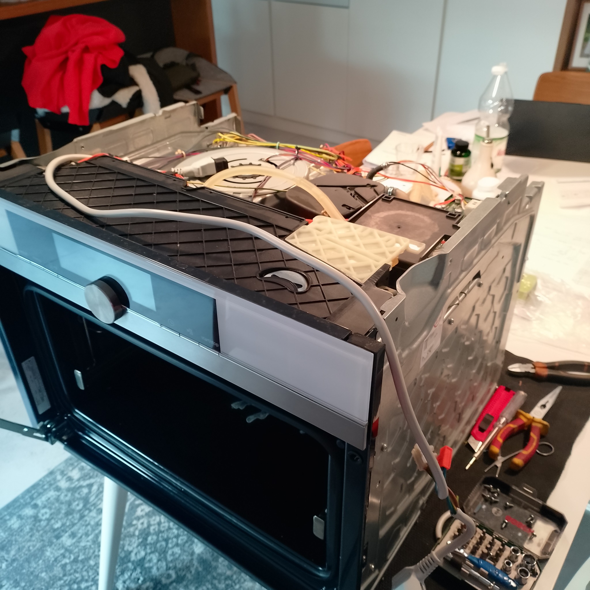 Rozłożona zmywarka na stole z widocznymi narzędziami, przewodami i elementami wewnętrznymi, sugerująca etap naprawy lub serwisu urządzenia.