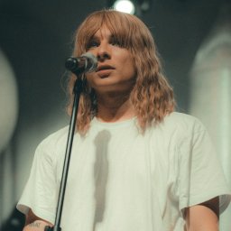 Paweł Woliński - Młody mężczyzna z długimi blond włosami w białej koszulce śpiewa do mikrofonu na scenie, widoczny tatuaż na ramieniu.