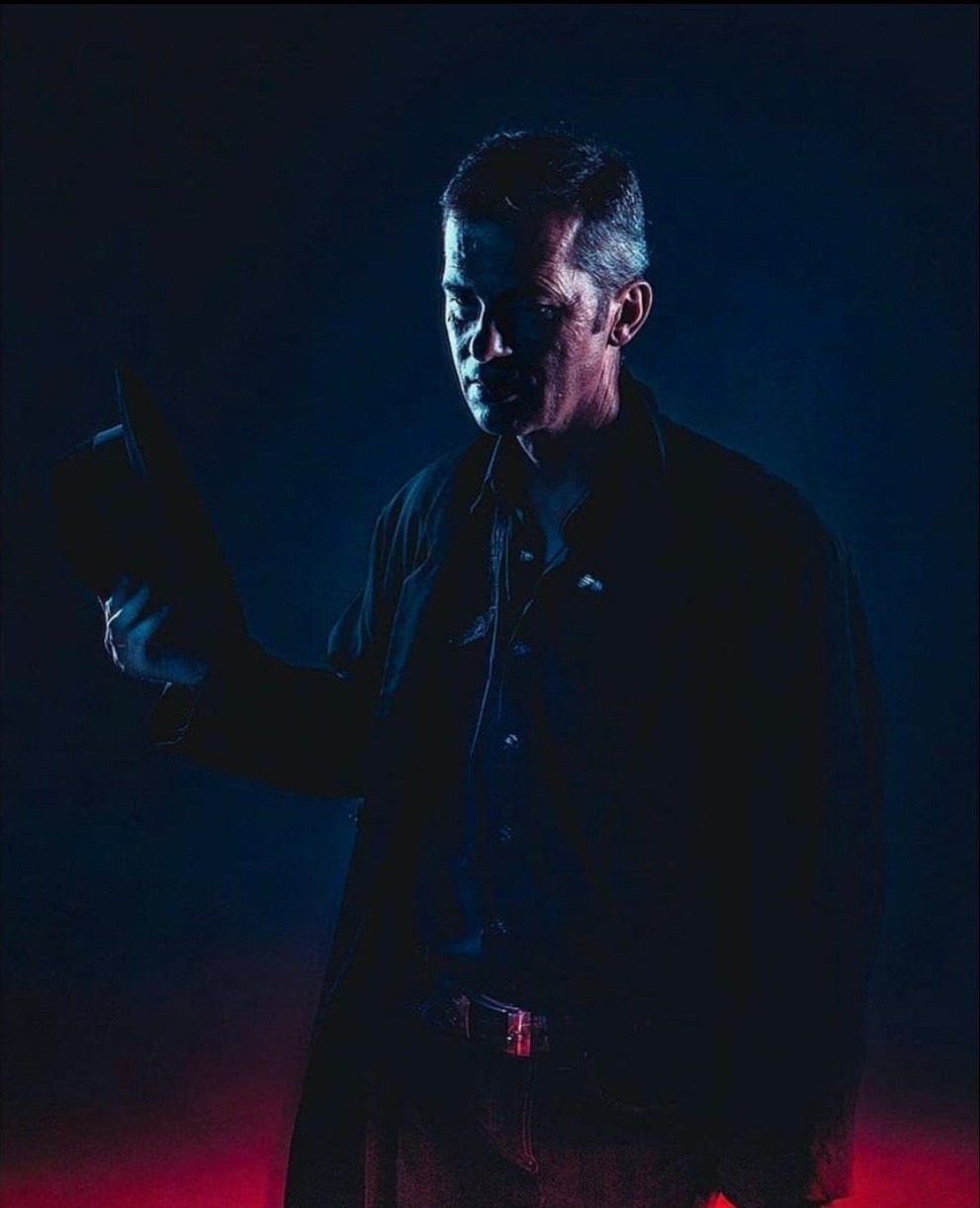 Mężczyzna w ciemnym ubraniu i kapeluszu w dłoni, oświetlony niebieskim i czerwonym światłem, tworzący dramatyczny portret.