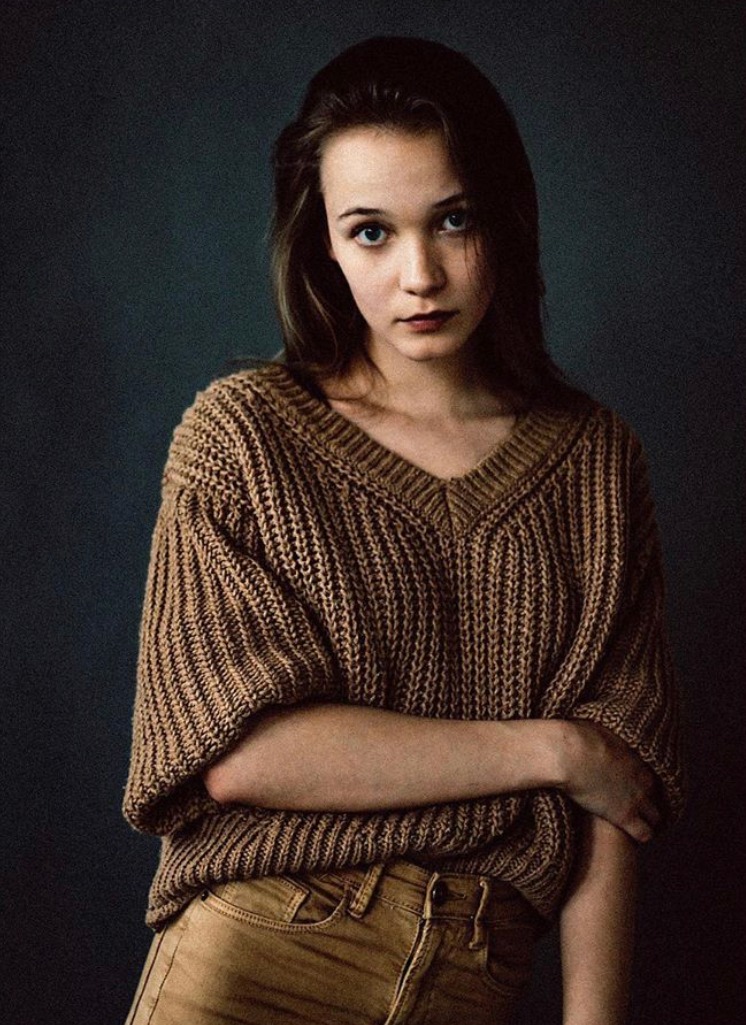 Portret młodej kobiety z niebieskimi oczami, ubrana w brązowy sweter z grubym splotem i spodnie w podobnym odcieniu na ciemnym tle.