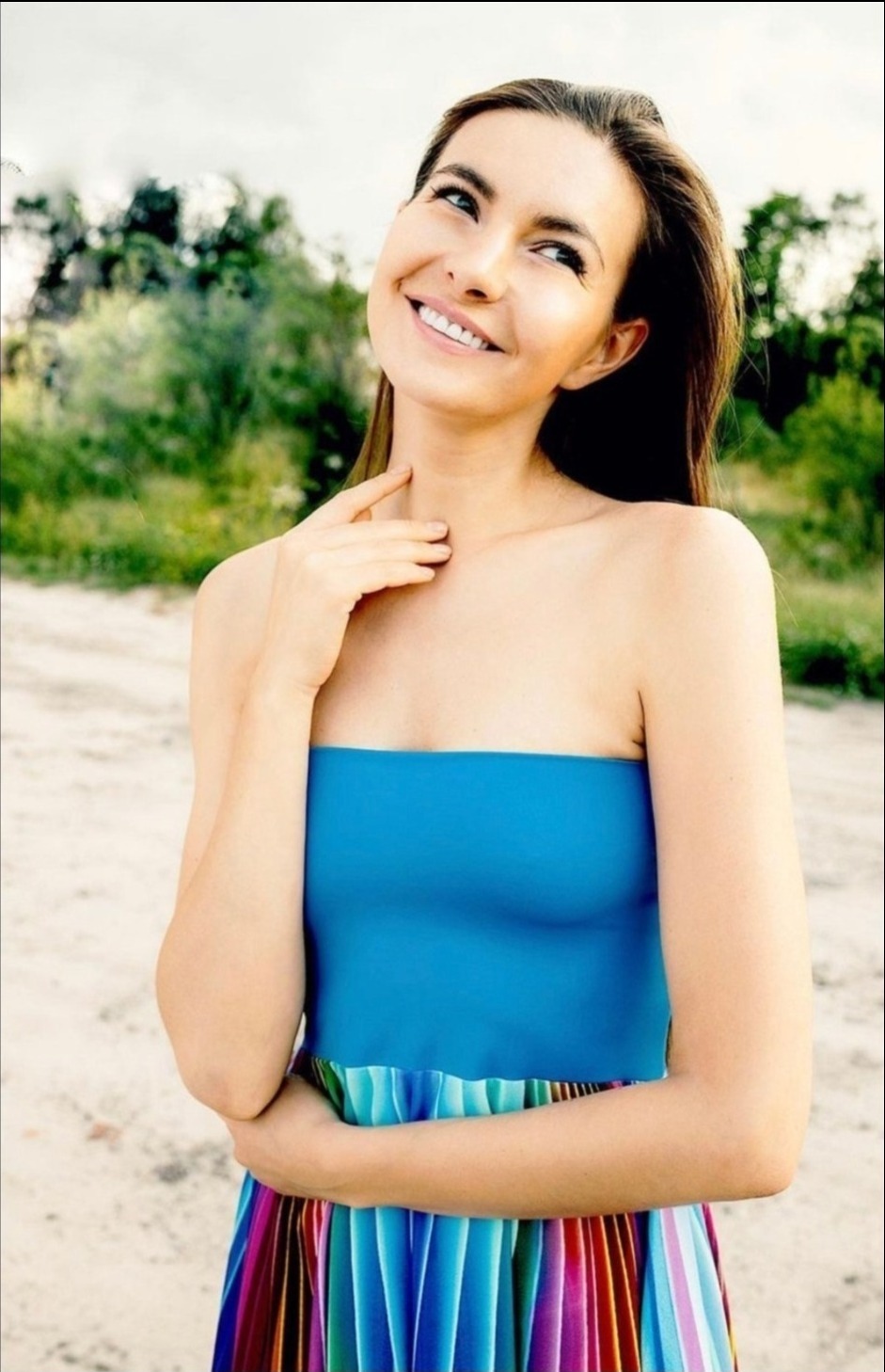 Uśmiechnięta kobieta w błękitnej sukience bez ramiączek, z kolorową plisowaną spódnicą, pozuje na tle zieleni, delikatnie dotykając szyi.