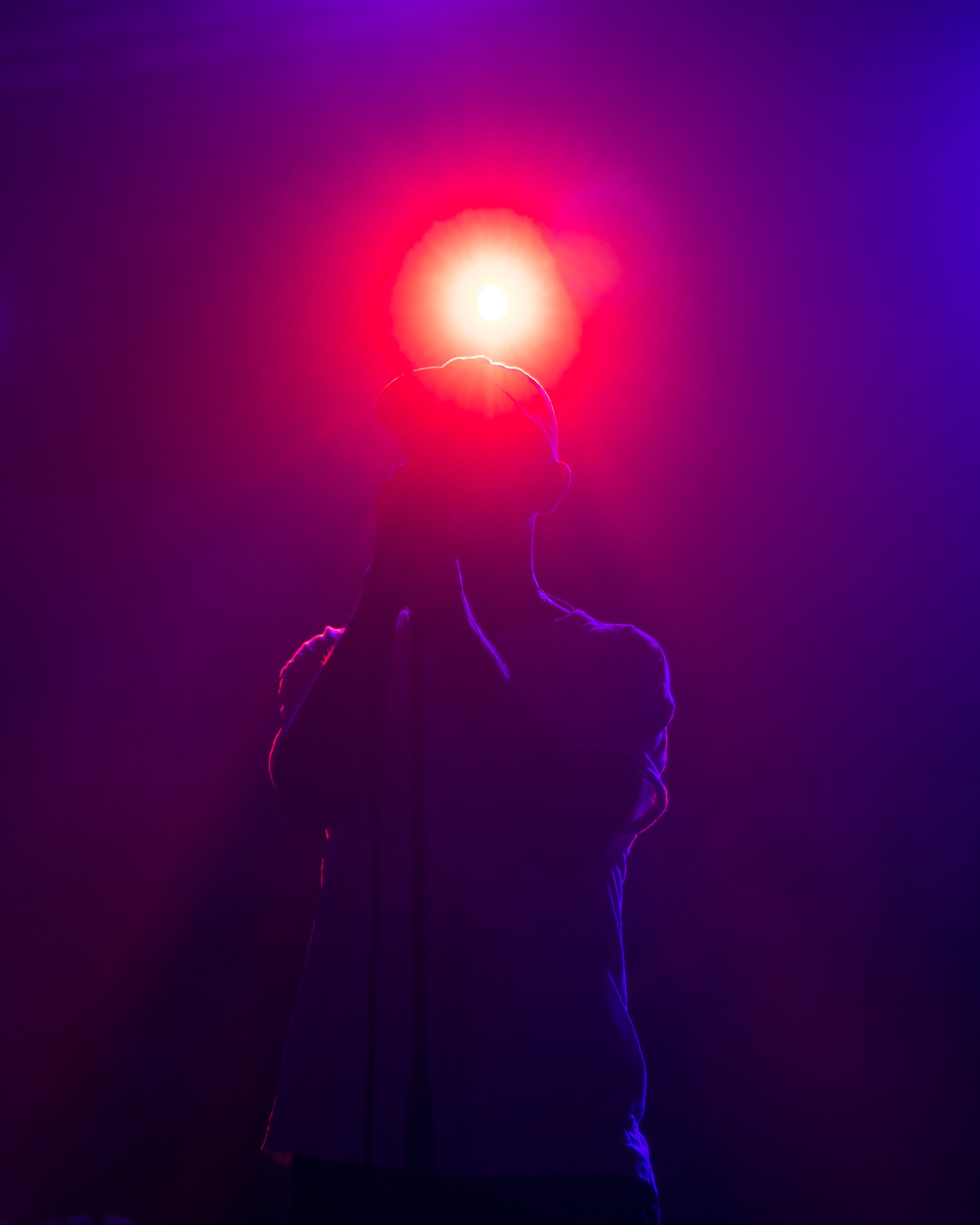 Sylwetka piosenkarza z mikrofonem na scenie oświetlonej czerwono-fioletowymi światłami.