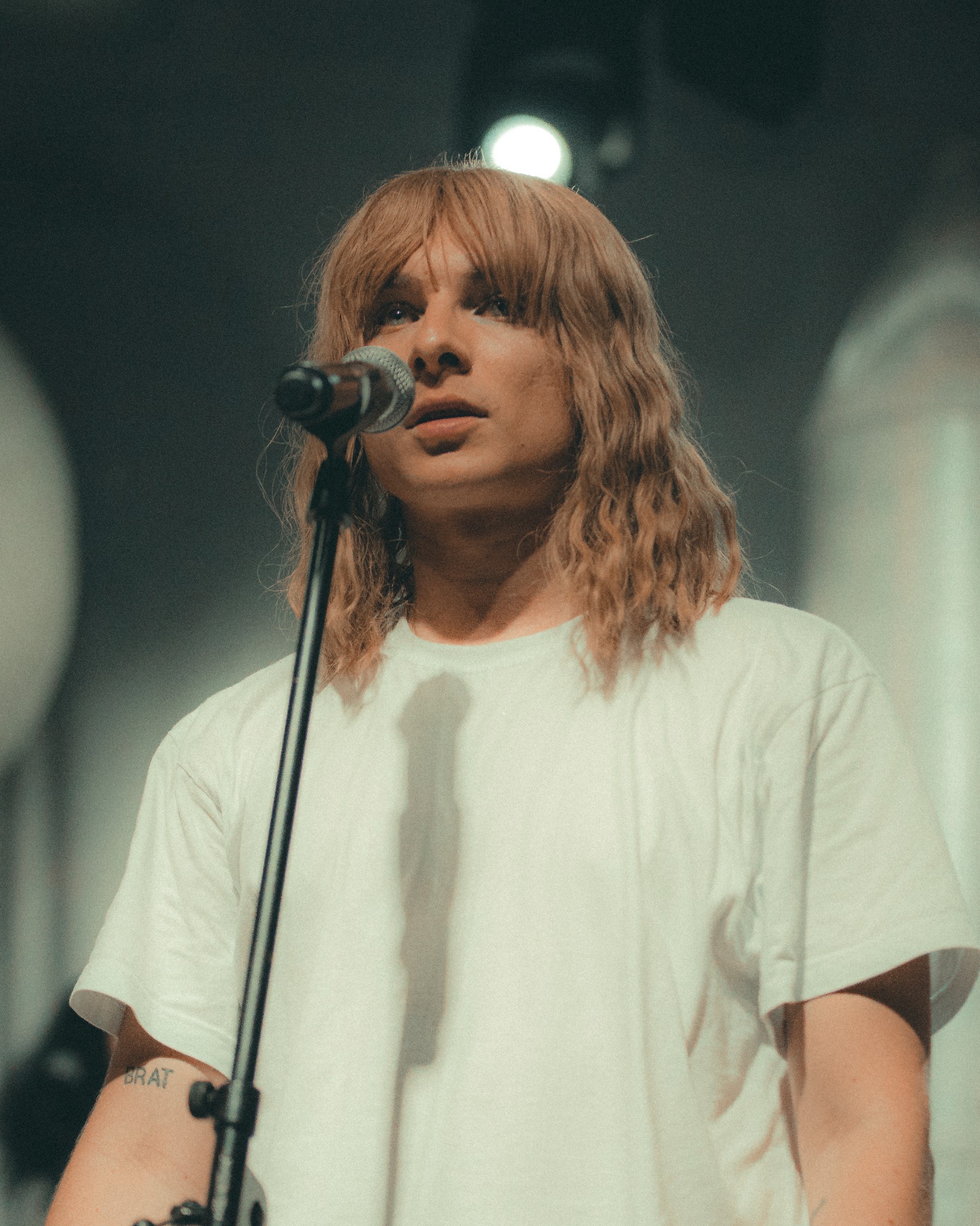 Młody mężczyzna z długimi blond włosami w białej koszulce śpiewa do mikrofonu na scenie, widoczny tatuaż na ramieniu.