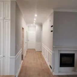 Wykończony korytarz w szaro-białej kolorystyce z drewnopodobną podłogą, zabudowaną szafą, ozdobnymi panelami ściennymi, kominkiem elektrycznym i oświetleniem punktowym.