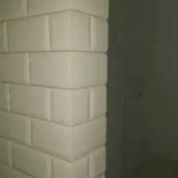 Wykończony narożnik ściany wyłożony białymi, prostokątnymi płytkami typu metro, widoczna szklana kabina prysznicowa w tle.