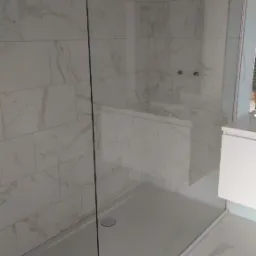 Kabina prysznicowa typu walk-in z białym brodzikiem i marmurowymi płytkami ściennymi, widoczne przyłącza wodne w tle.