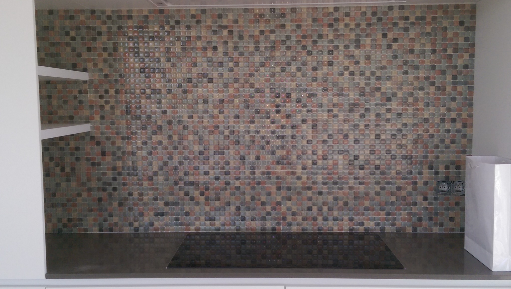 Mozaika z małych, kwadratowych płytek w odcieniach brązu, szarości i beżu, tworząca backsplash nad ciemnym blatem kuchennym z płytą indukcyjną; widoczne białe półki i gniazdka elektryczne.