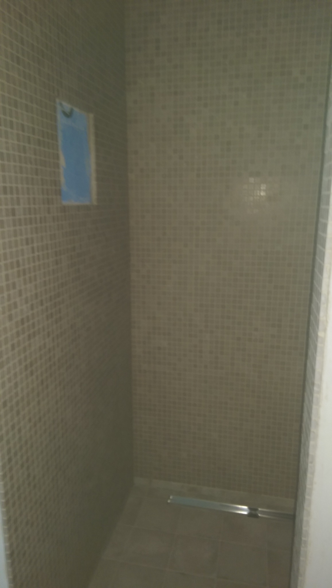 Wnętrze kabiny prysznicowej wyłożonej mozaiką w odcieniach beżu i szarości, z widocznym odpływem liniowym i fragmentem zabezpieczonej ściany przed położeniem płytek.