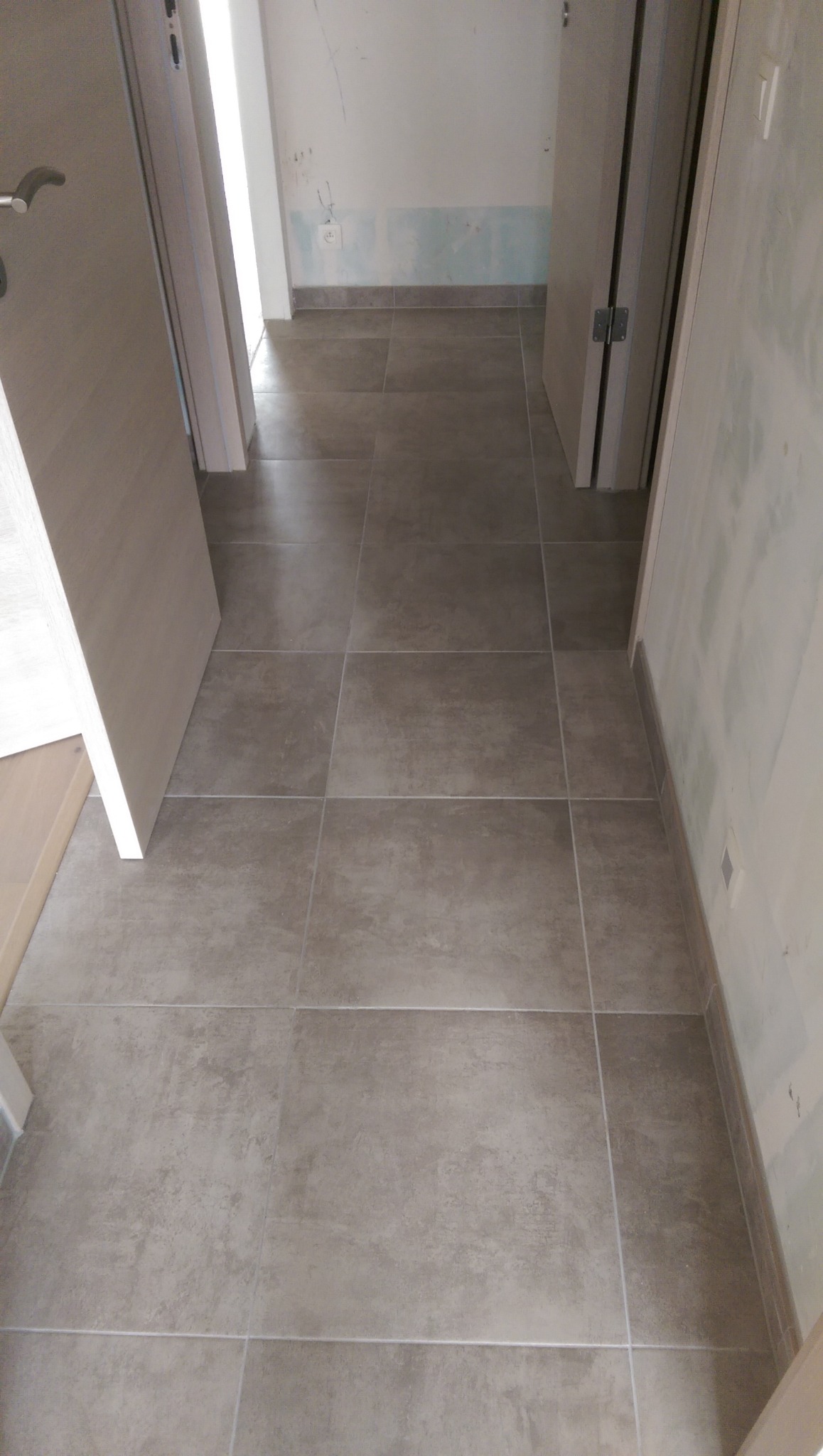 Ułożone szare płytki podłogowe w korytarzu, widoczne fragmenty ścian z śladami po remoncie i otwarte drzwi.