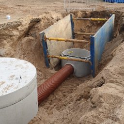 Selbud Sebastian Staszewski - Wykop zabezpieczony stalowymi szalunkami, widoczny betonowy zbiornik z brązową rurą karbowaną, w tle koparka i baraki budowlane.