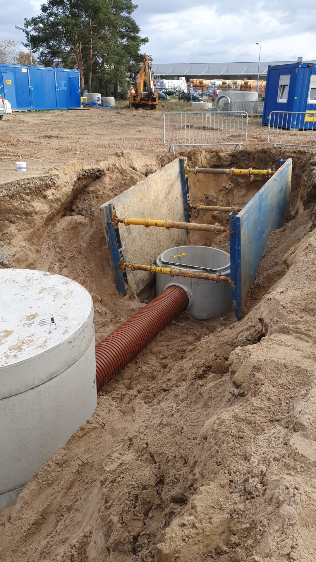 Wykop zabezpieczony stalowymi szalunkami, widoczny betonowy zbiornik z brązową rurą karbowaną, w tle koparka i baraki budowlane.