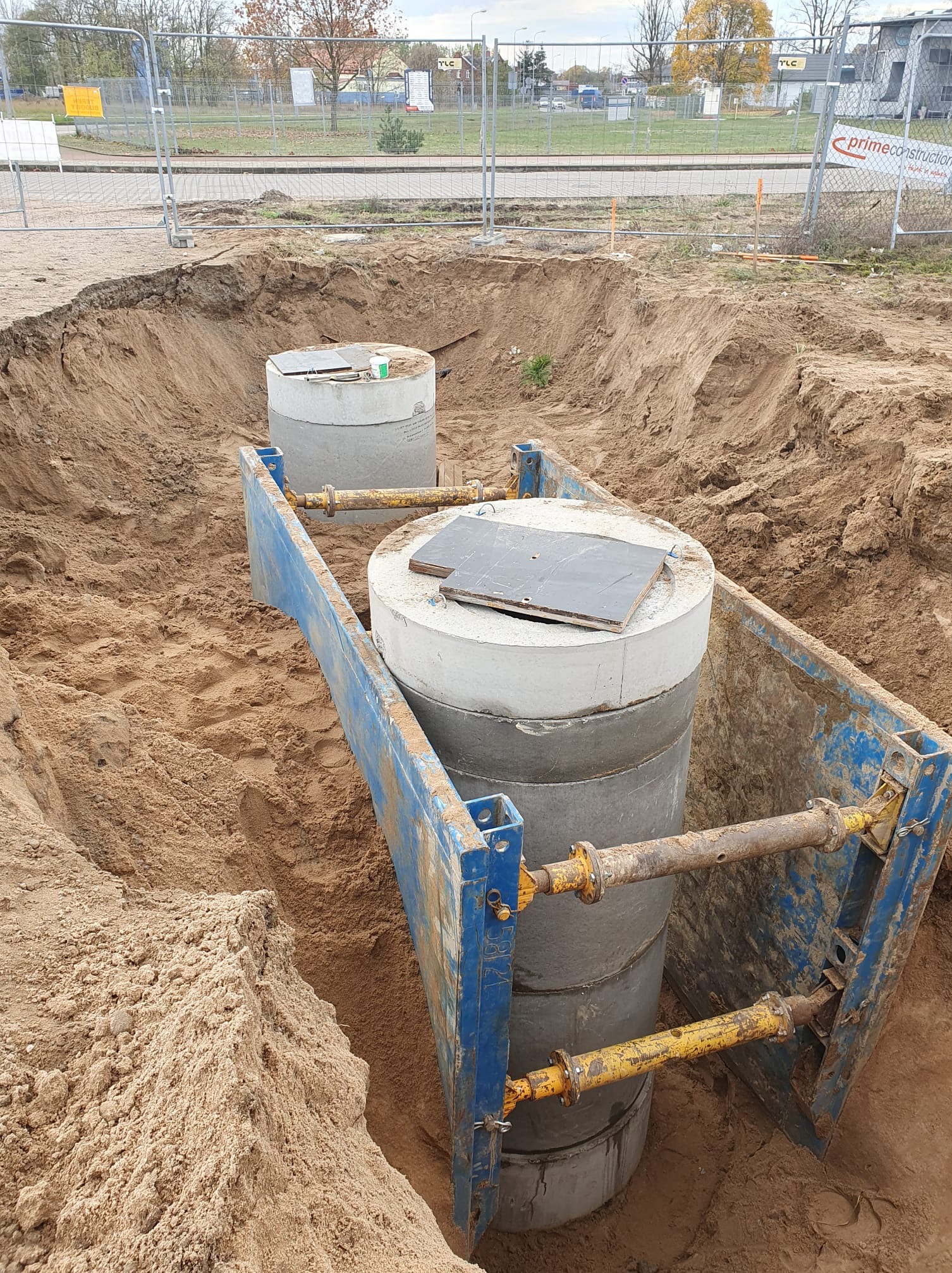 Dwa betonowe kręgi w wykopie zabezpieczonym niebieską szalunką, widoczna warstwa piasku i ziemi, na górnym kręgu leży płyta, w tle ogrodzenie i budynek.