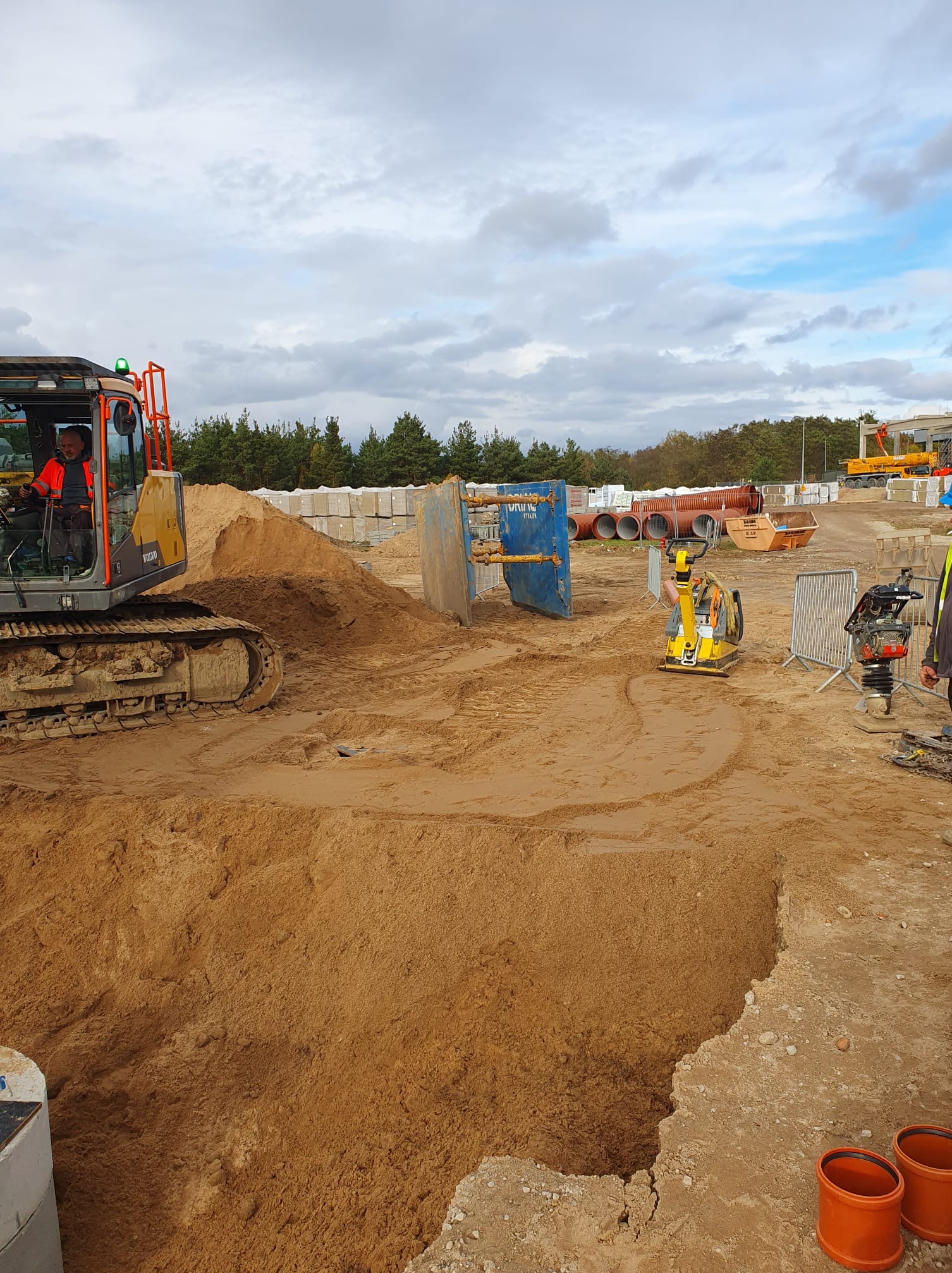 Wykonawca w kabinie koparki Volvo, przygotowuje wykop w piasku, widoczne pomarańczowe rury, zagęszczarki i zabezpieczenie wykopu.