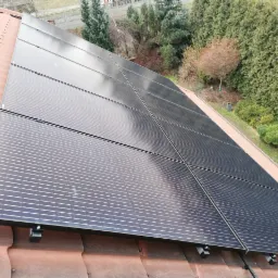 Panele słoneczne zamontowane na dachu z czerwonej dachówki, widok z góry pod kątem, w tle drzewa i krzewy.