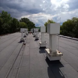 Widok z góry na płaski dach pokryty papą z rzędem białych, kwadratowych kominów wentylacyjnych, z widocznymi przewodami i elementami instalacji, na tle zachmurzonego nieba i zieleni drzew.