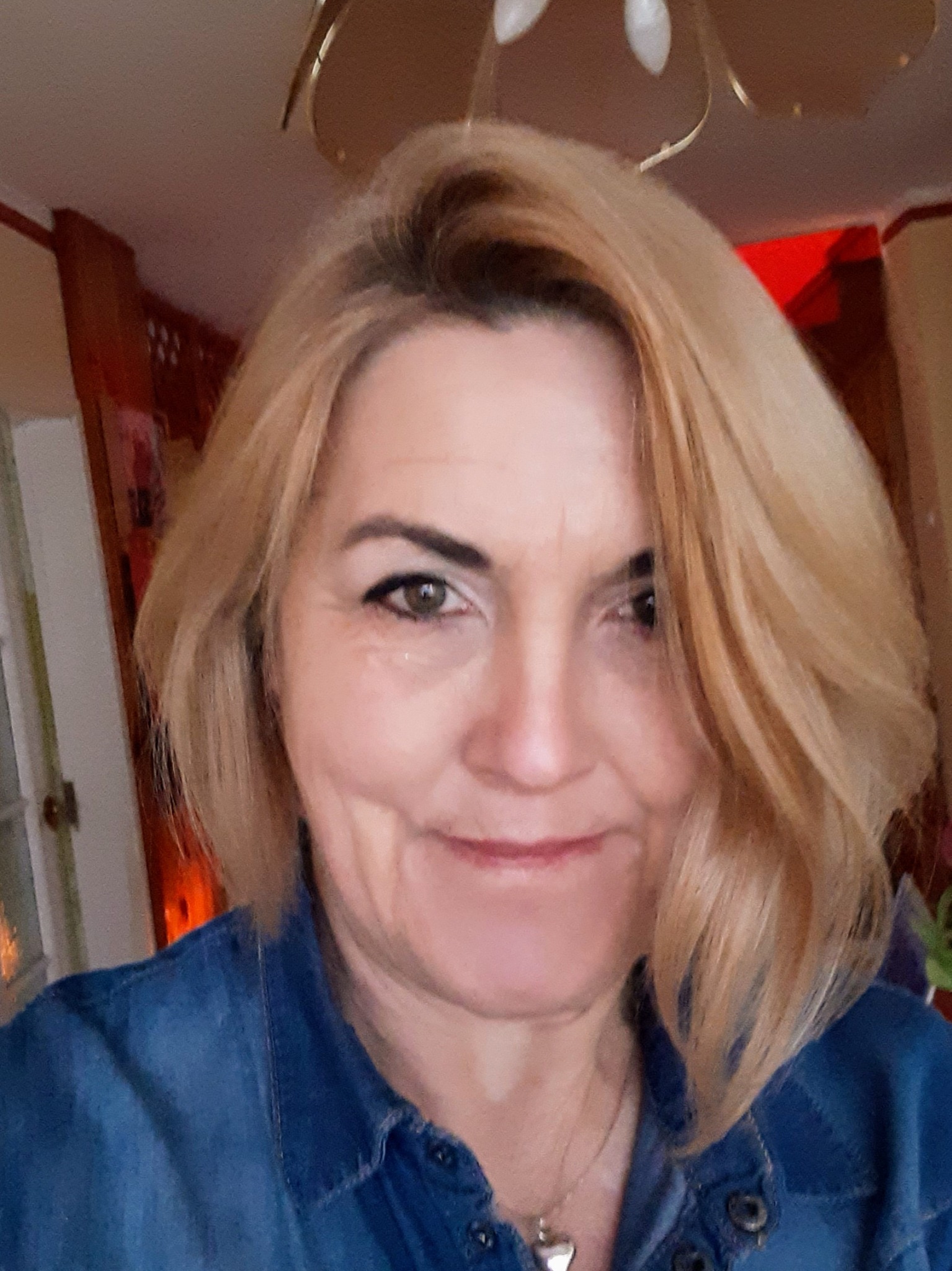 Portret uśmiechniętej kobiety z blond włosami w denimowej koszuli, z widocznym fragmentem żyrandola i drzwi w tle.