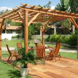 Drewniana altana z prostą konstrukcją kratową, z wiszącymi roślinami, pod którą znajduje się stół z krzesłami na drewnianym tarasie, otoczona zielonym trawnikiem i roślinnością.