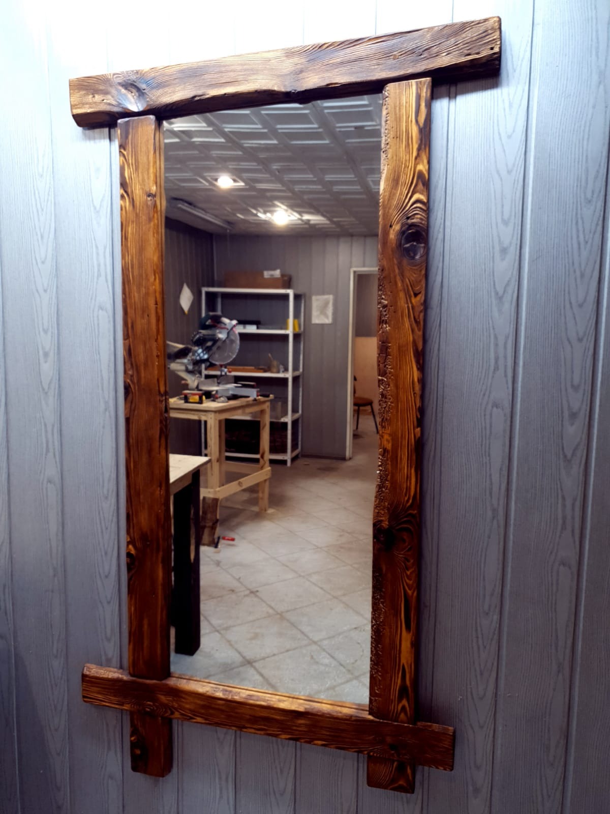 Ręcznie wykonane lustro z ramą z belek sosnowych, widoczne sęki i unikalna struktura drewna, odbicie warsztatu w tafli lustra, szare panele ścienne w tle.