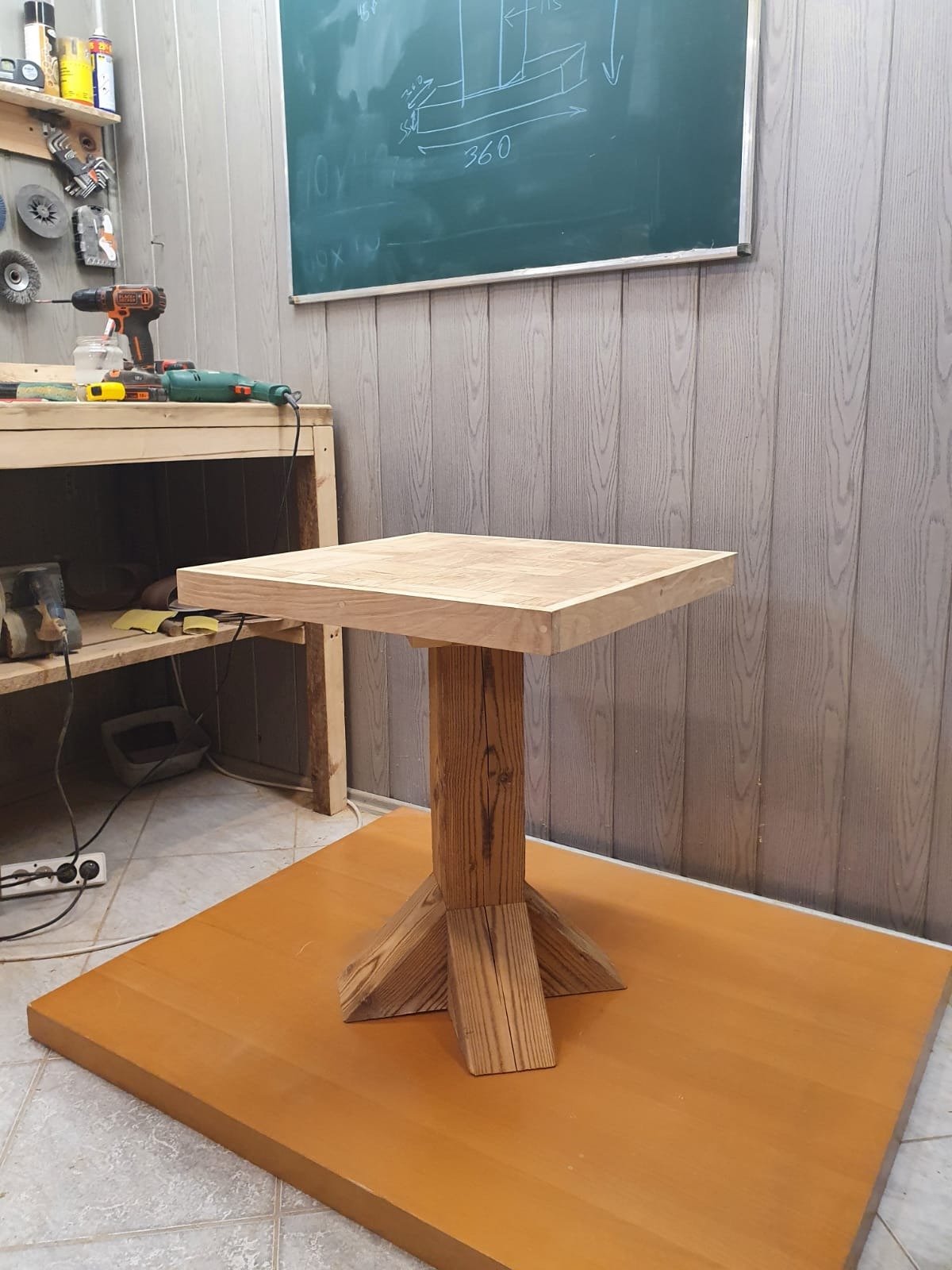 Surowy, kwadratowy stolik kawowy z dębowym blatem i sosnową podstawą w warsztacie, widoczna tablica z rysunkiem technicznym w tle.